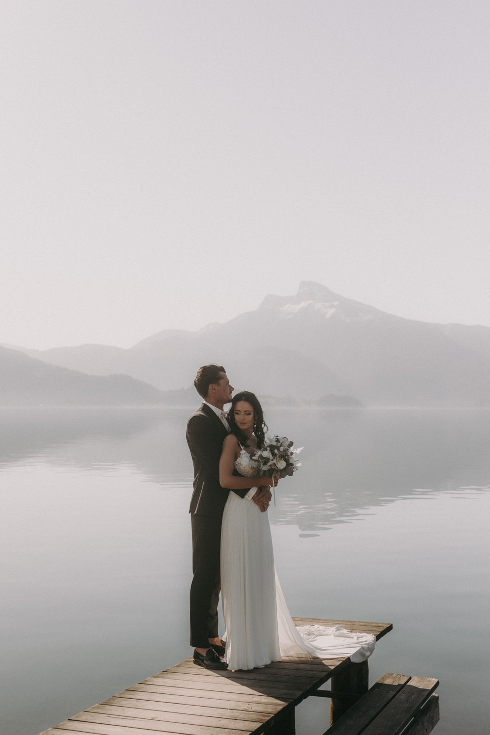 Hochzeitspaar in liebevoller Pose auf Steg vor Alpenpanorama