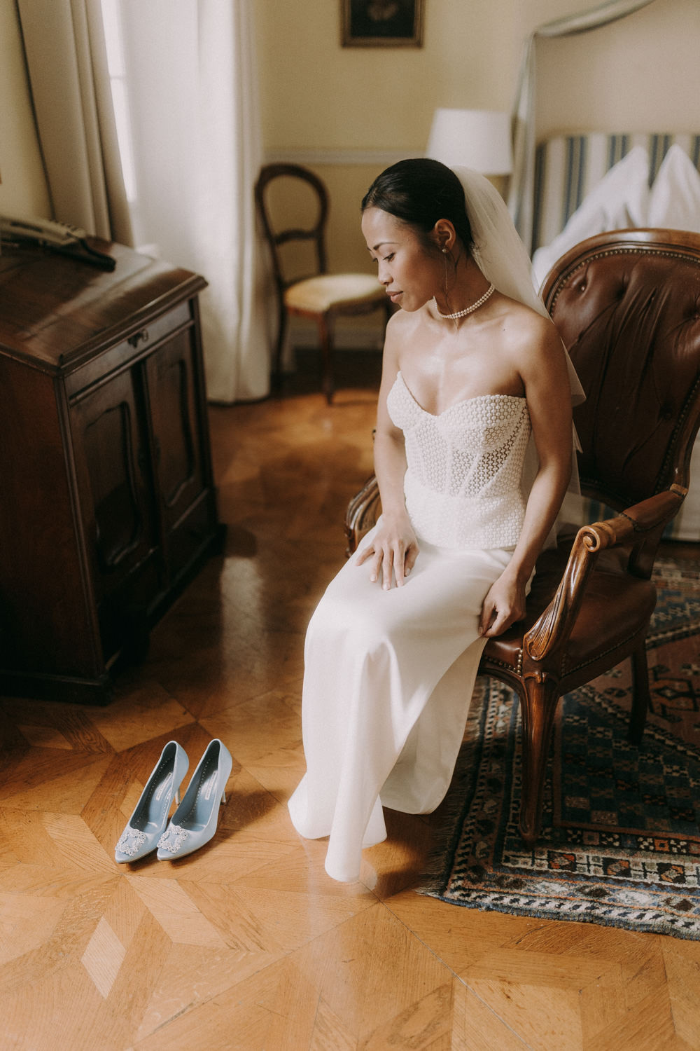 Bride in pearl-textured Kaviar Gauche gown seated indoors beside blue Manolo Blahnik shoes.