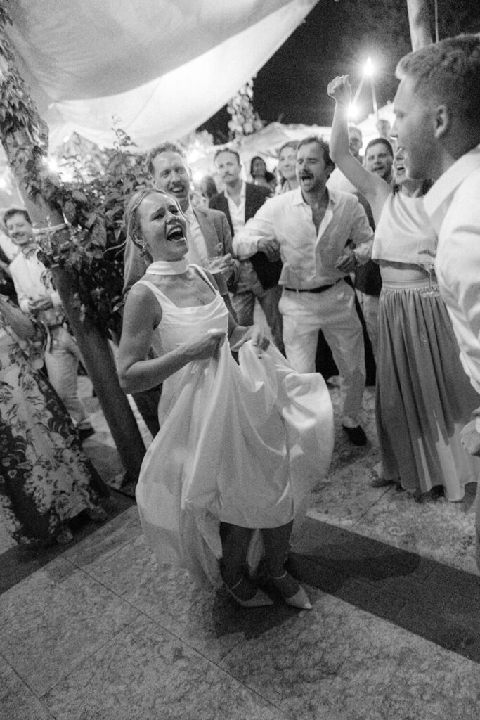 Bride laughing and dancing surrounded by cheering guests under string lights, black and white flash photography.