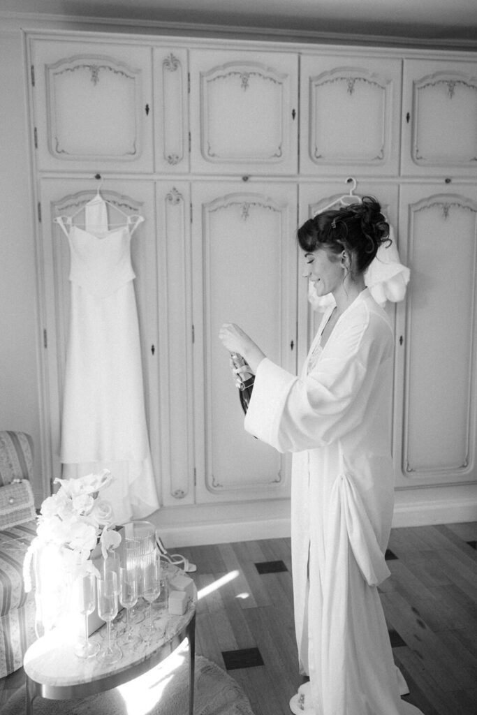 Bride in a robe opening a bottle of champagne next to her hanging wedding dress, black and white getting ready moment.