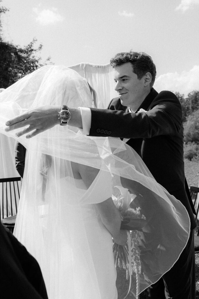 Groom reaching out to embrace his bride as her veil billows in the wind after the ceremony, black and white wedding photography.