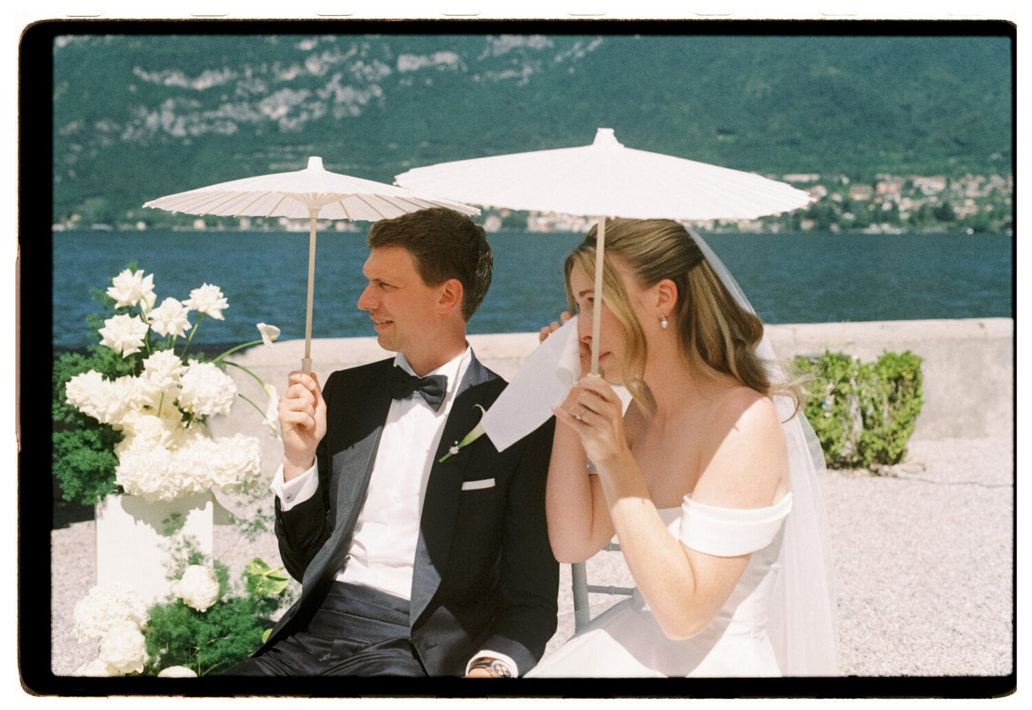 Bride wiping away a tear under a white paper parasol beside her groom during their lakeside ceremony on Lake Como, shot on 35mm film