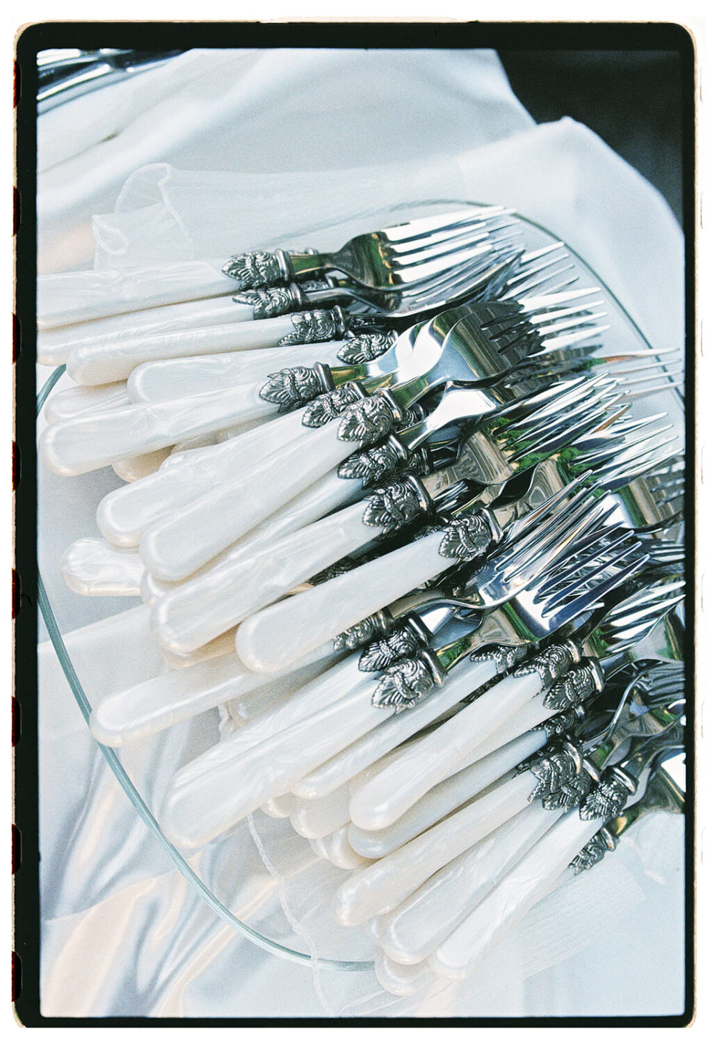 Pearl-handled forks with ornate silver caps stacked on a glass plate over white linen, wedding detail on 35mm film.