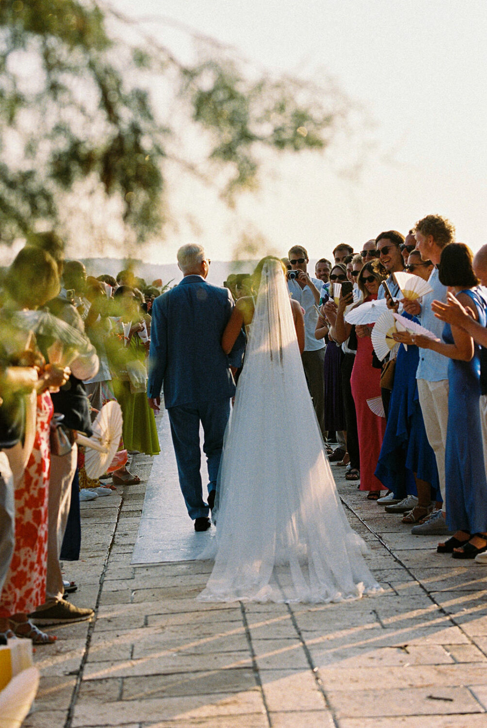 ride in a long veil walks down the aisle on her father’s arm between guests with paper fans and parasols, Sicily wedding on 35mm film