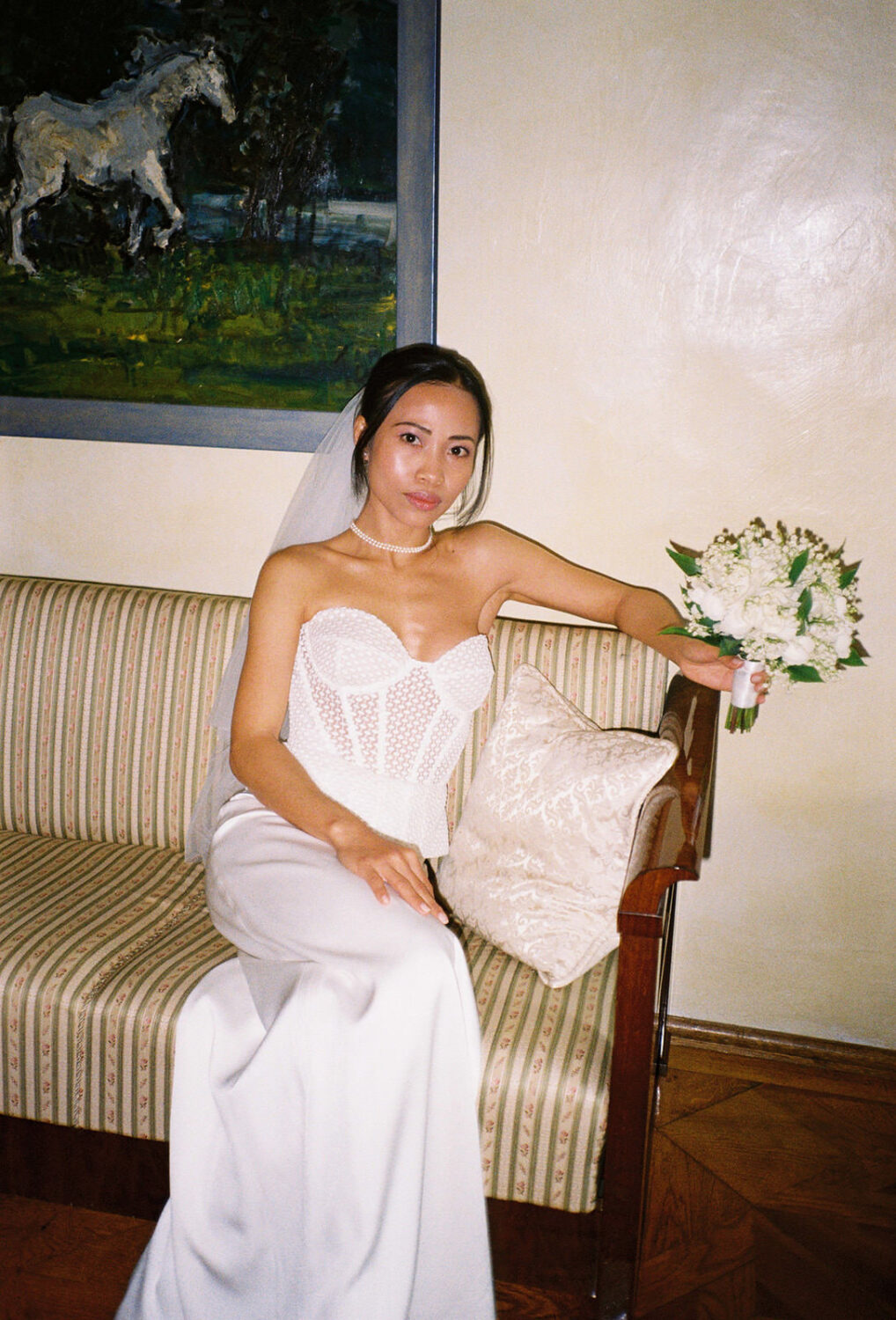 Bride in a lace corset gown and veil sitting on a striped antique sofa in a Tuscan villa, holding a white bouquet, shot on 35mm film
