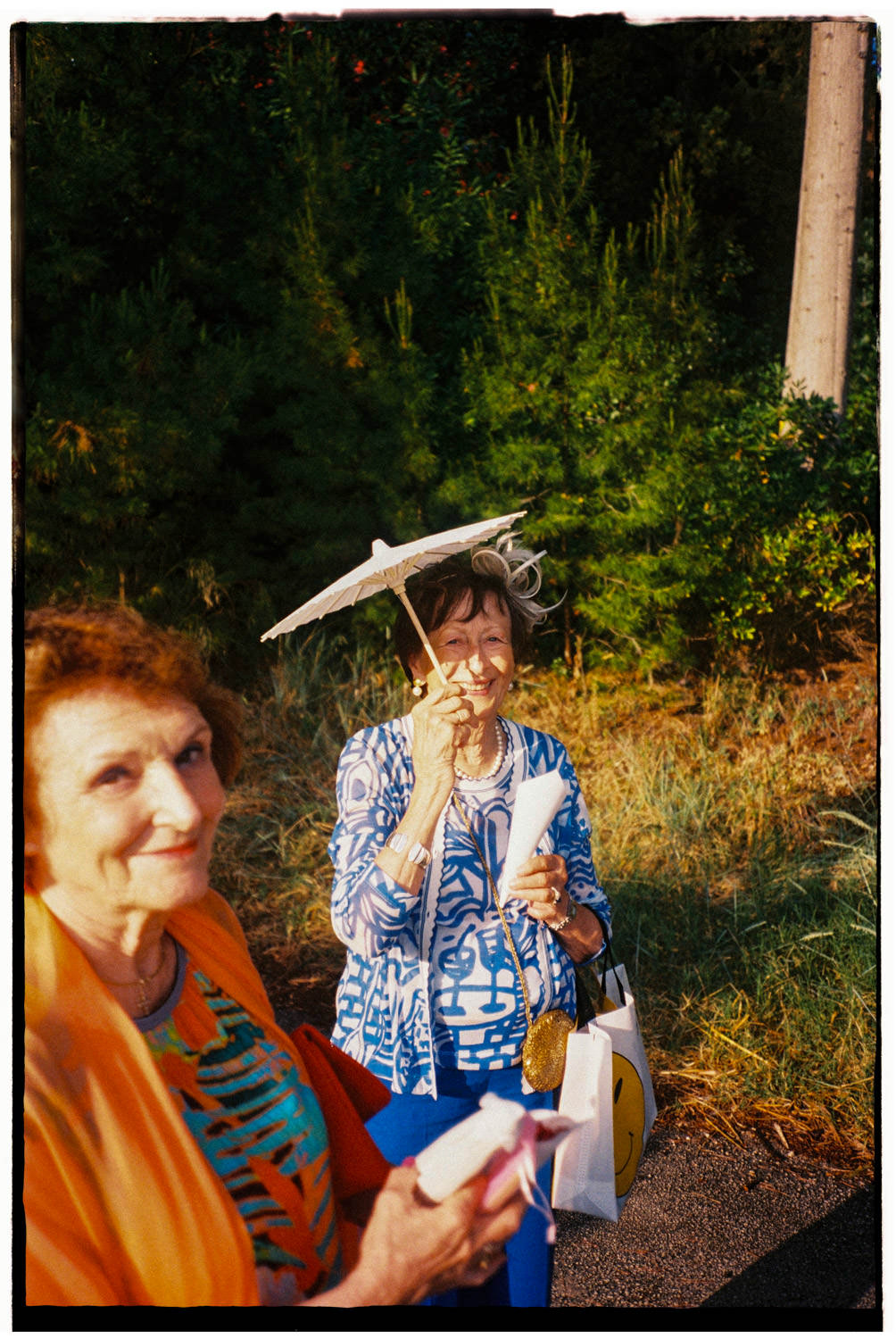 Two elegant older wedding guests smiling in the golden evening sun at Villa Dalmacija in Split, one holding a white paper parasol, shot on 35mm film.